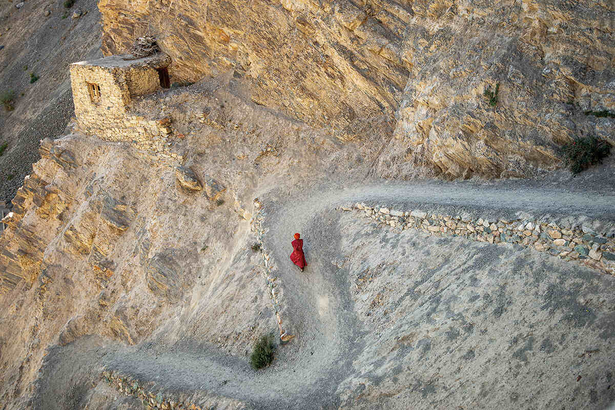 A monk in red robes walks along a mountainous dirt path to a monastary