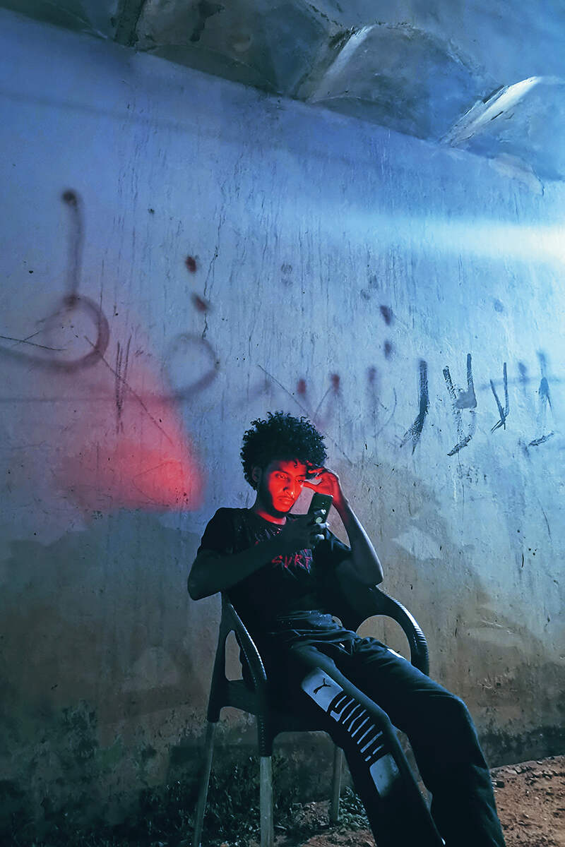 A man sitting on a chair in a dark alleyway illuminated by red light and blue light
