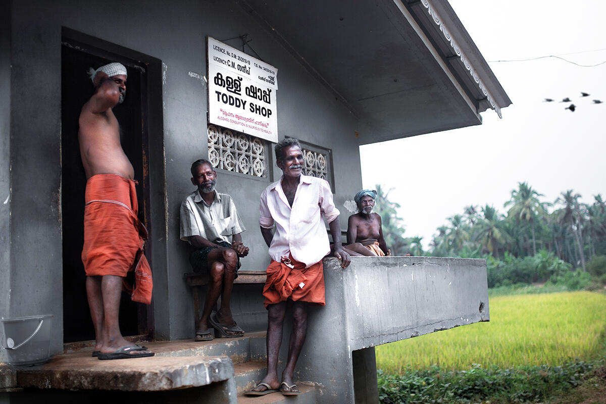 A group of indian men stand outside a grey 'toddy shop'