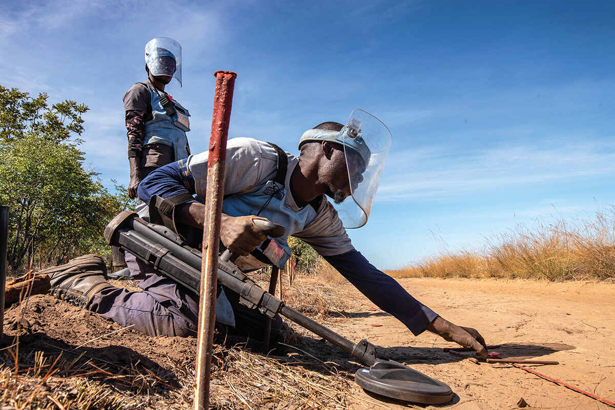 A man on his knees marks the location of a landmine in the dirt with some sticks