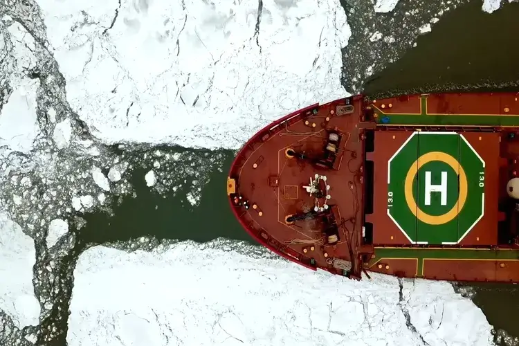 Aerial top-down view of a powerful icebreaker ship cutting through thick frozen sea ice in the Arctic waters of Finland. The vessel creates a narrow path through the white icy surface, revealing dark green water beneath. The striking contrast between the red ship deck, marked helipad, and the surrounding frozen landscape highlights navigation, exploration, and the harsh beauty of polar environments.