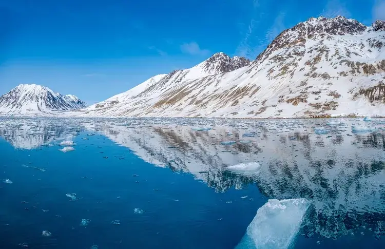 Breathtaking Arctic landscape in Svalbard, Norway, featuring snow-capped mountains reflecting in the crystal clear water of Magdalenefjord with floating ice floes on a sunny day.