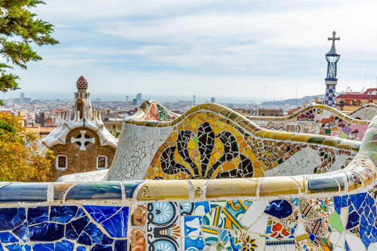 Colorful mosaics on fence of serpentine bench in main square below the Hypostyle of Parc Guell, urban cityscape against cloudy sky in background, sunny day in Barcelona, Catalonia, Spain