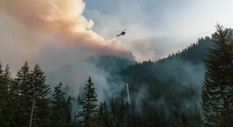 A firefighting helicopter drops water over a smoke-filled conifer forest during a wildfire — Smoke from Wildfires near Hope, BC Canada