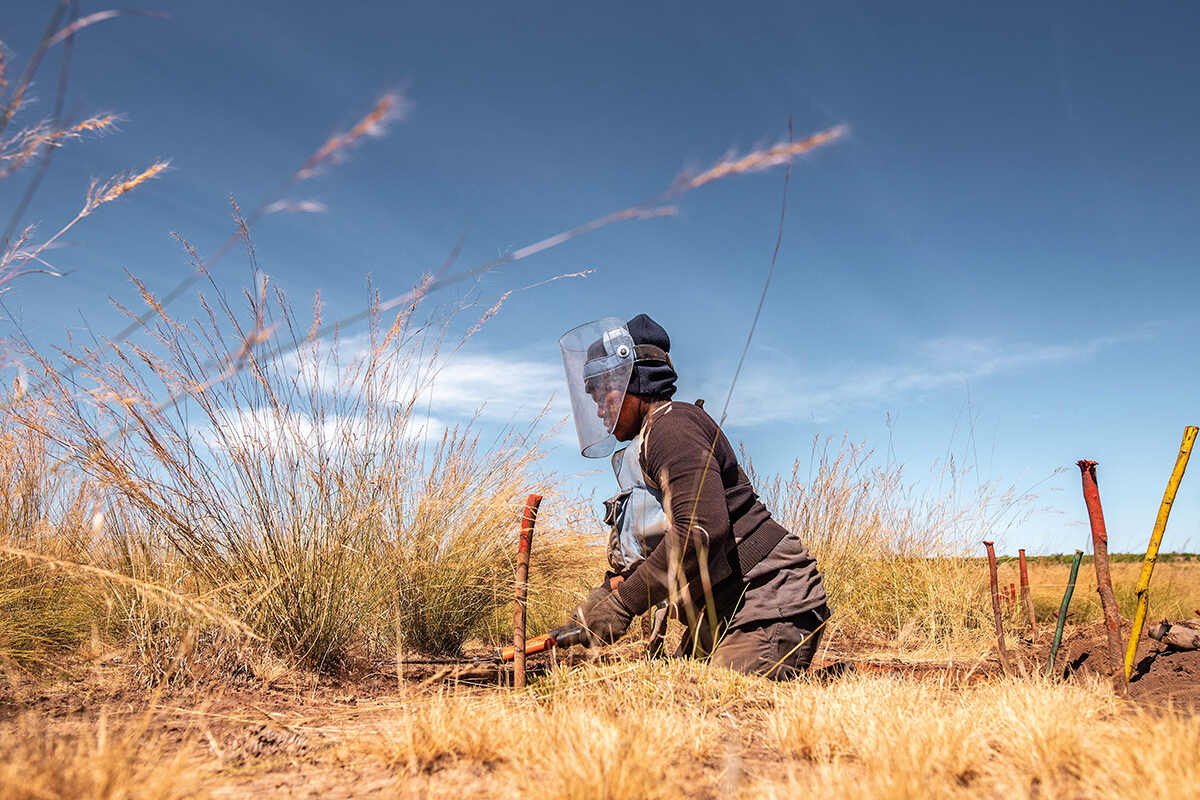 A woman on her knees cutting back vegetation in search of landmines