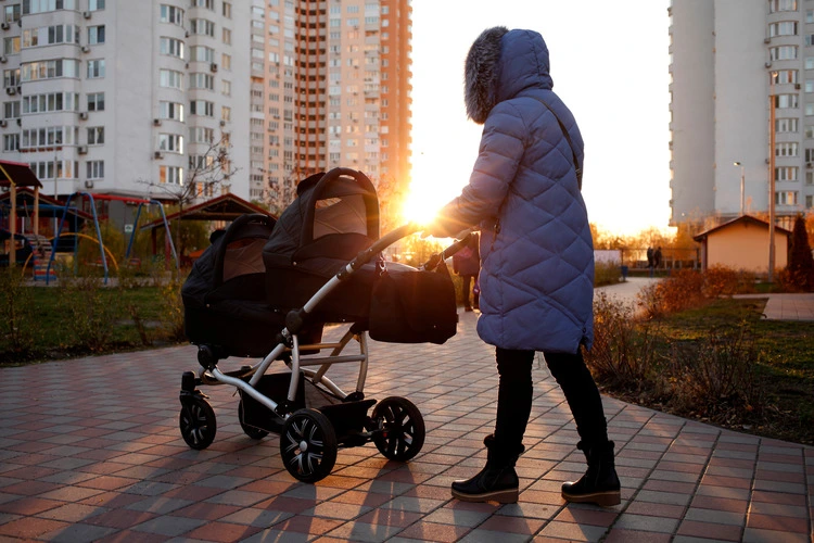 Young mother walking in a park with children in pushchair. Mom and kids in a buggy walk in forest. Woman pushing a double stroller for twin boy newborn baby. Active family outdoors.