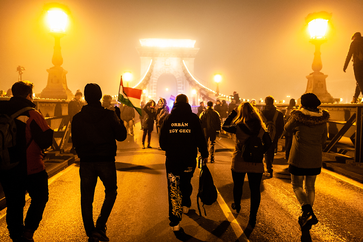 A group of people march over a bridge in Hungary during a protest at night, orange hue