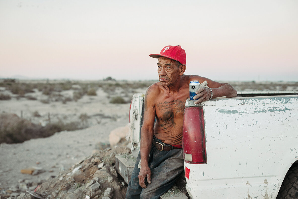 A shirtless man sits drinking a beer on the bed of a truck