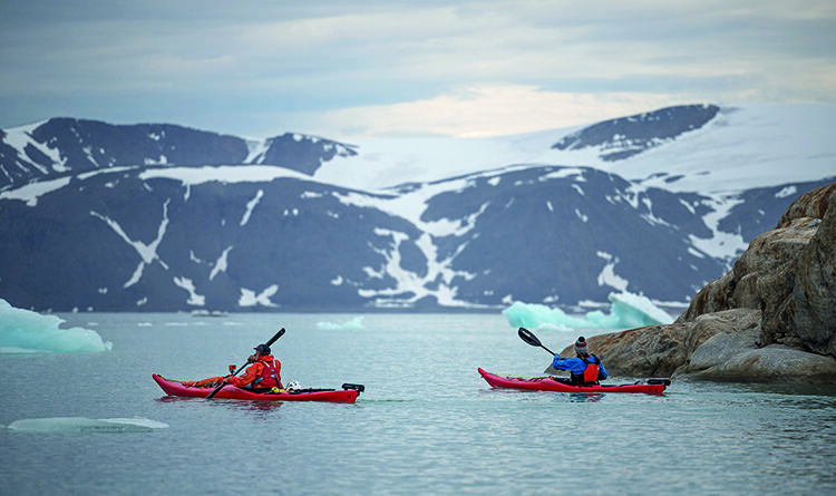 Two kayakers paddle between rocls and icebergs on East Greenland's coast