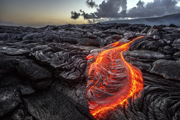 Red-orange vibrant molten lava flowing onto grey lava field and glossy rocky land near Hawaiian volcano