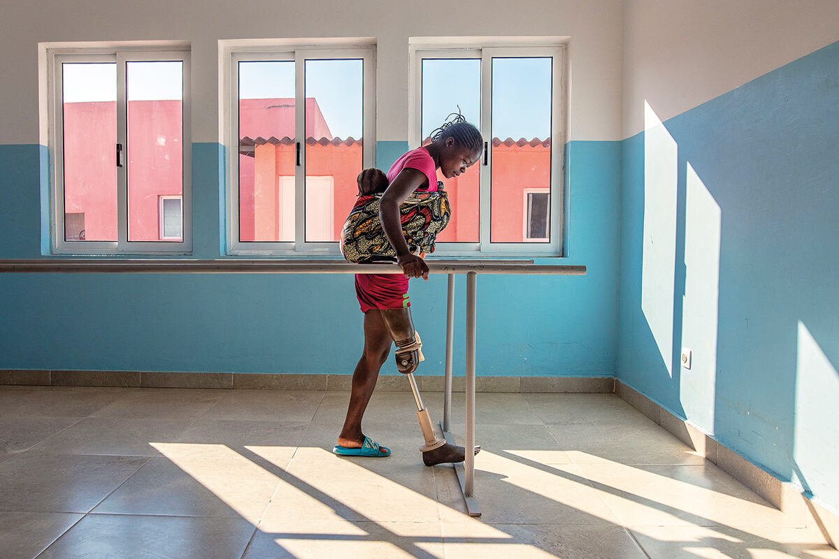 A woman in a bare room carrying a baby on her back and holding onto rails learns to walk again on a prosthetic