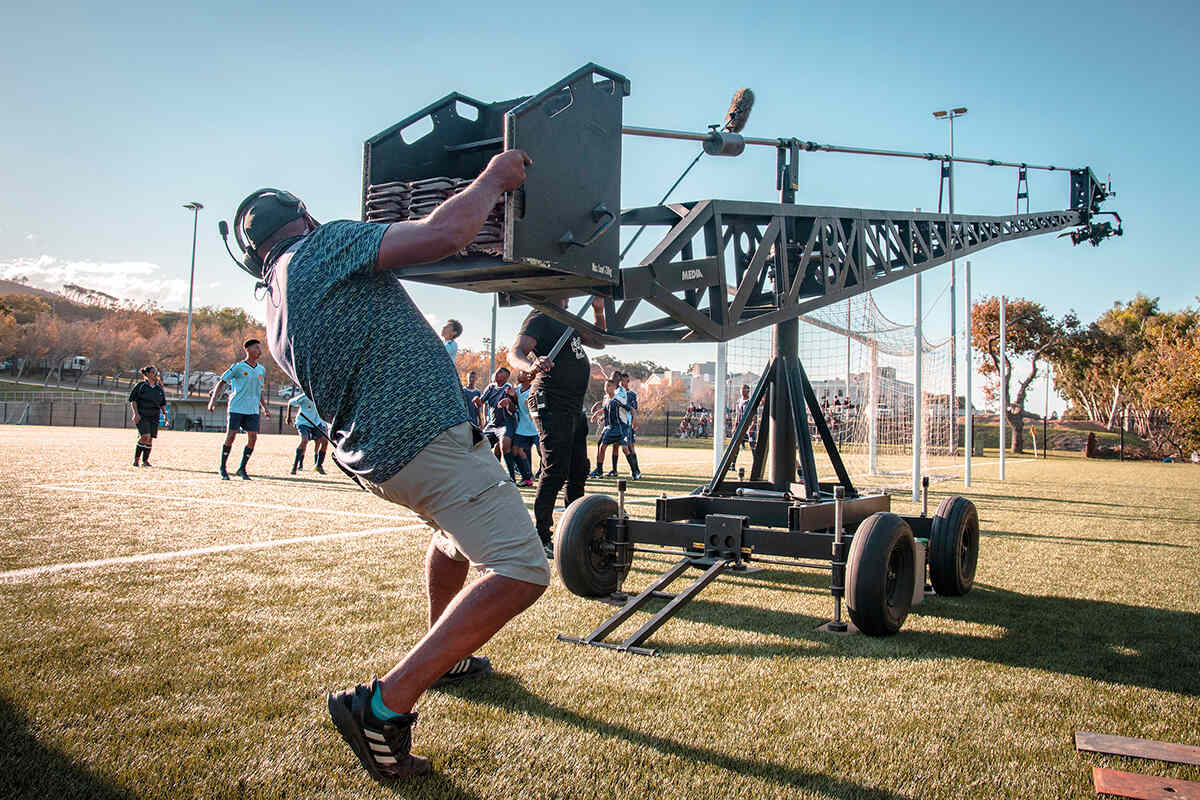 A man handles an overhead camera crane while shooting a scene with footballers on a pitch