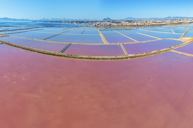 Mar Menor aerial view with pink & blue gradient water, with view of mountains and skyline in background.