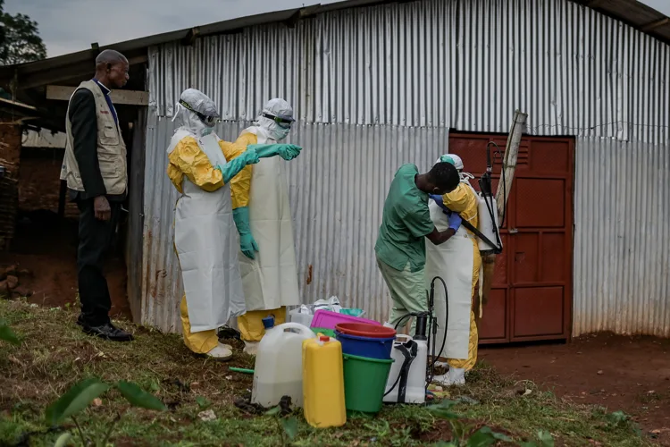 A priest beside health workers in protective equipment outside a metal-walled building in Mongbwalu