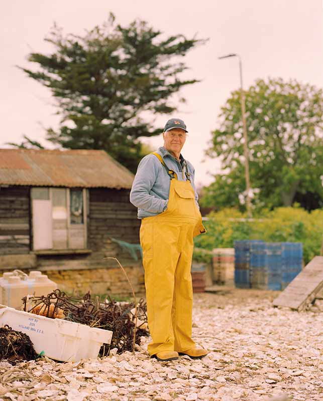 An oysterman in yellow overalls stands on oyster shells