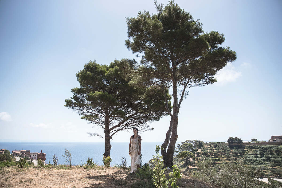 A woman standing between two trees on a cliff overlooking the sea performs a shoiw