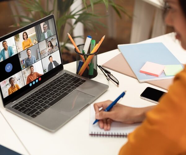 A woman sits at a desk with a laptop open, displaying a video conference meeting with several participants. She is taking notes in a notebook with a blue pen, her focus on the screen.