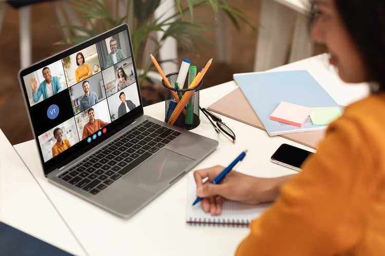 A woman sits at a desk with a laptop open, displaying a video conference meeting with several participants. She is taking notes in a notebook with a blue pen, her focus on the screen.