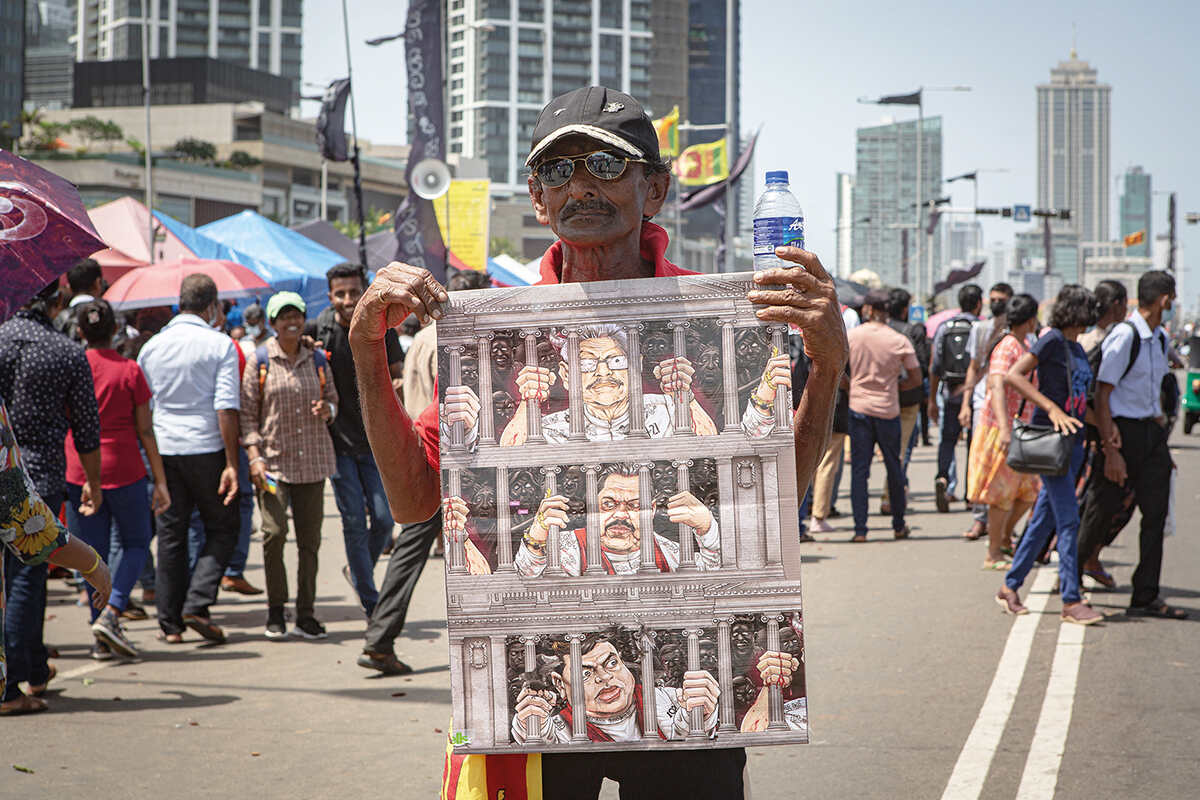 A man stands in a street protest holding a cartoon poster of the Rajapaska Family