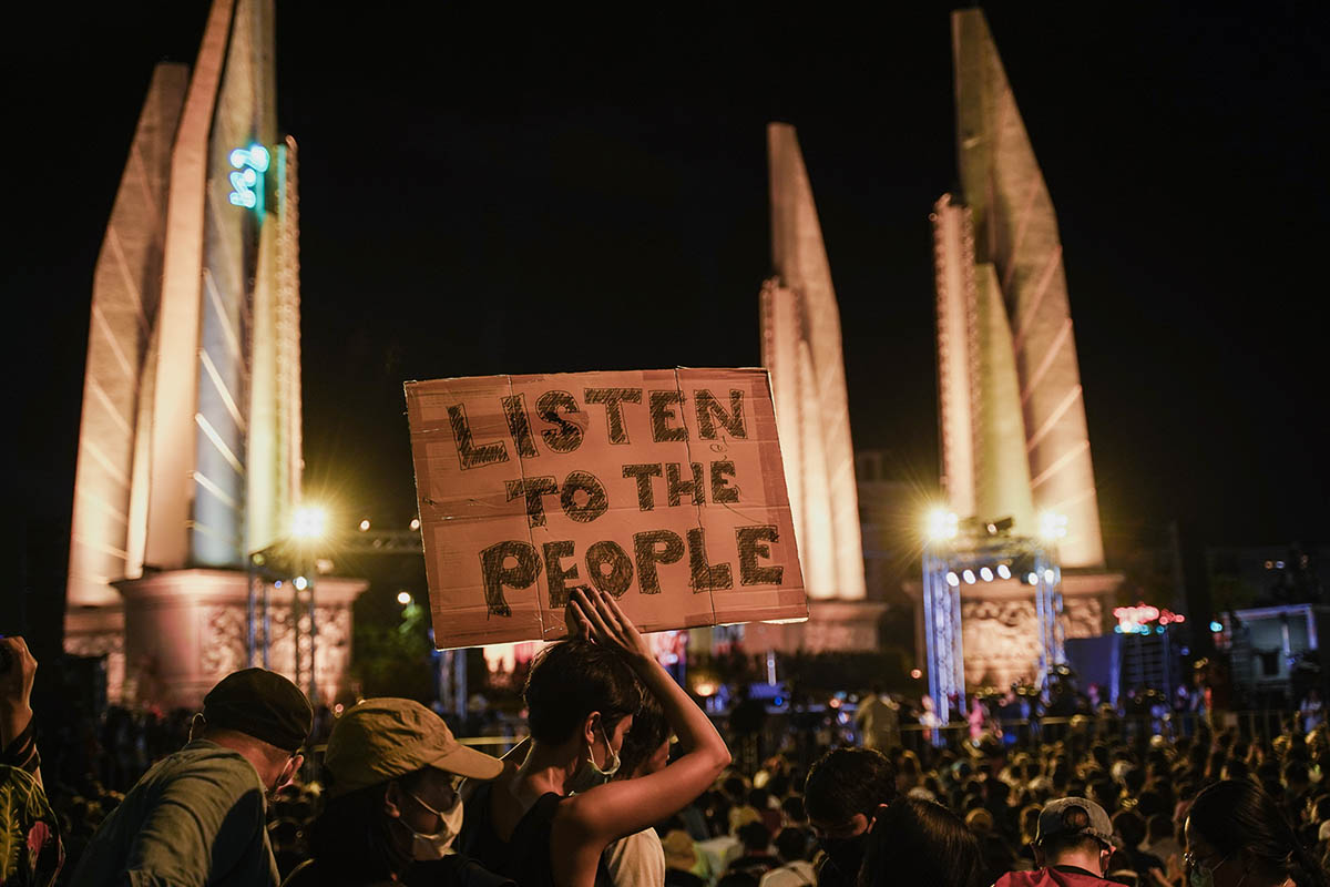 A woman holds up a cardboard sign reading 'listen to the people' during a protest at night