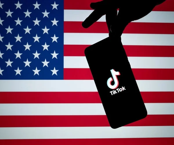 TikTok logo seen on the silhouette of smartphone hold in a hand with American flag on the back.