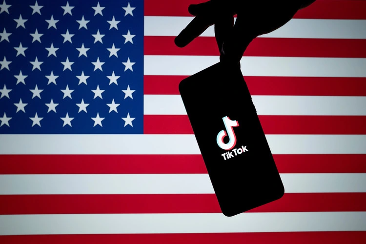 TikTok logo seen on the silhouette of smartphone hold in a hand with American flag on the back.