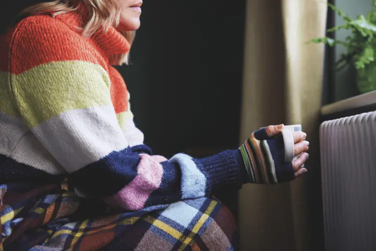 A woman holds a mug with a long-sleeve bright coloured jumper, next to a radiator.