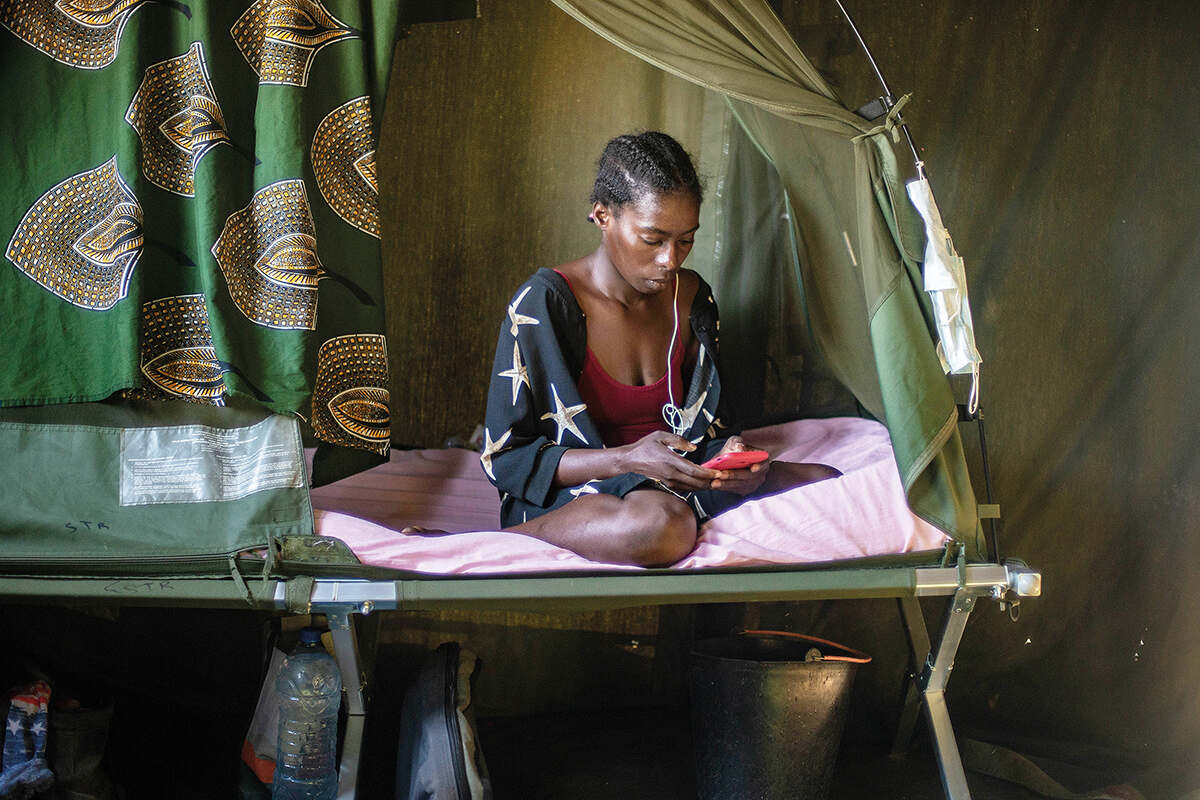 A woman listens to music through headphones on her phone will sitting on a camp bed in camp